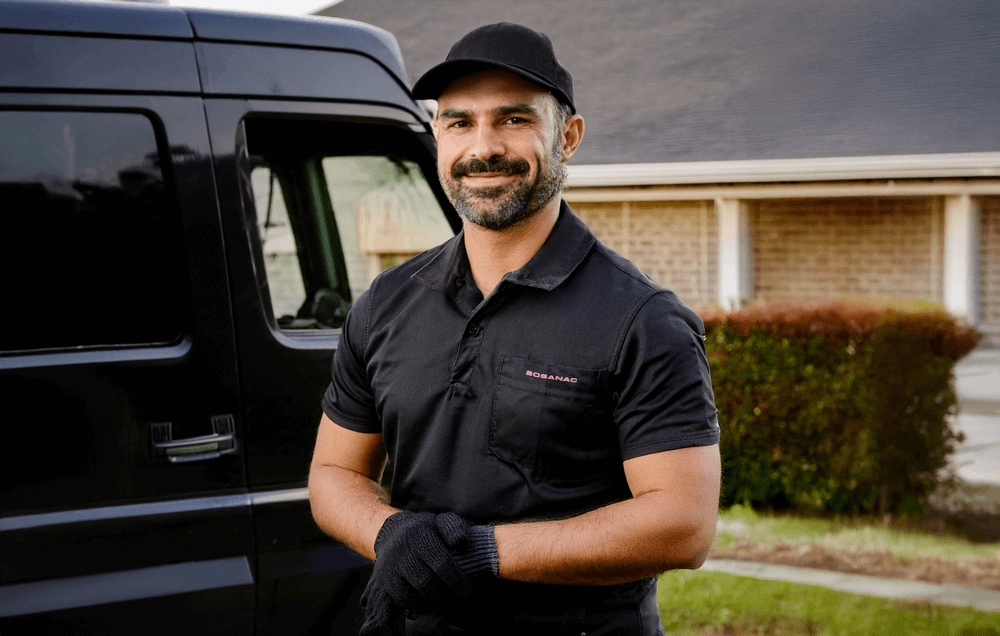 A smiling Bosanac technician stands outdoors beside a black service van, wearing a black polo shirt, black cap, and black gloves. A residential house with a bush is visible in the background. A smiling Bosanac technician stands outdoors beside a black service van, wearing a black polo shirt, black cap, and black gloves. A residential house with a bush is visible in the background.