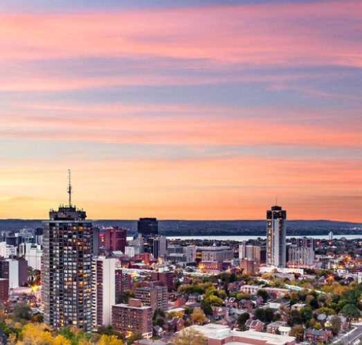 City skyline at sunset featuring two illuminated high-rise buildings and residential areas with a river in the background. City skyline at sunset featuring two illuminated high-rise buildings and residential areas with a river in the background.