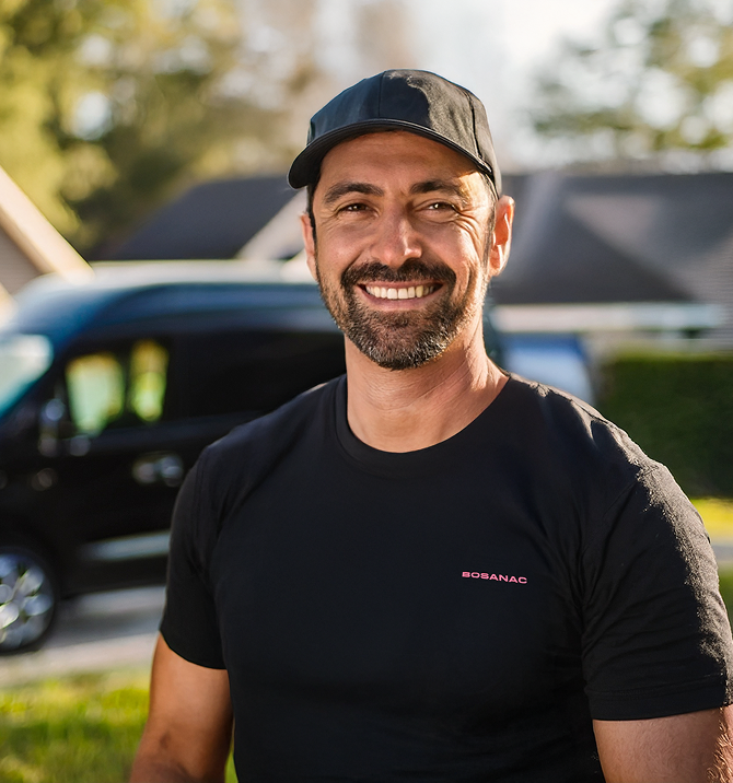 A smiling Bosanac crew member wearing a black cap and a black t-shirt with the Bosanac logo stands outdoors with a blurred background of a vehicle and houses in soft daylight. A smiling Bosanac crew member wearing a black cap and a black t-shirt with the Bosanac logo stands outdoors with a blurred background of a vehicle and houses in soft daylight.