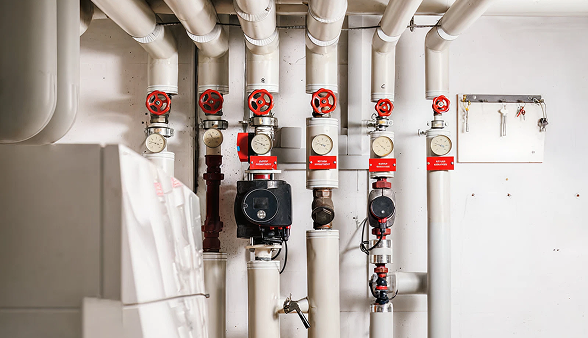 A Bosanac commercial industrial boiler system featuring white pipes, pressure gauges, and red valve handles installed on a white wall. A Bosanac commercial industrial boiler system featuring white pipes, pressure gauges, and red valve handles installed on a white wall.