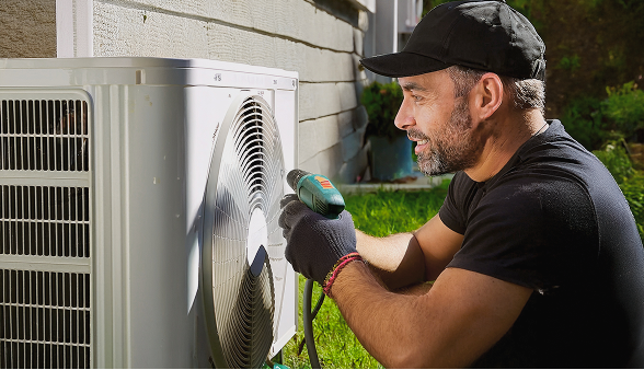 A Bosanac technician services an outdoor air conditioning unit with a handheld tool in a sunlit garden. A Bosanac technician services an outdoor air conditioning unit with a handheld tool in a sunlit garden.