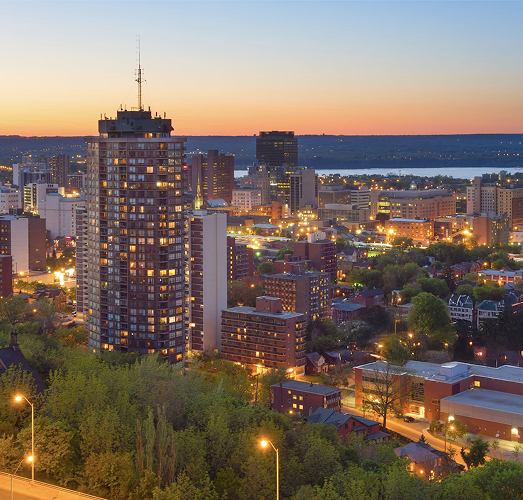 vening cityscape of Hamilton, featuring illuminated residential and commercial buildings, green tree-lined streets, and a distant river under a fading sunset sky. vening cityscape of Hamilton, featuring illuminated residential and commercial buildings, green tree-lined streets, and a distant river under a fading sunset sky.
